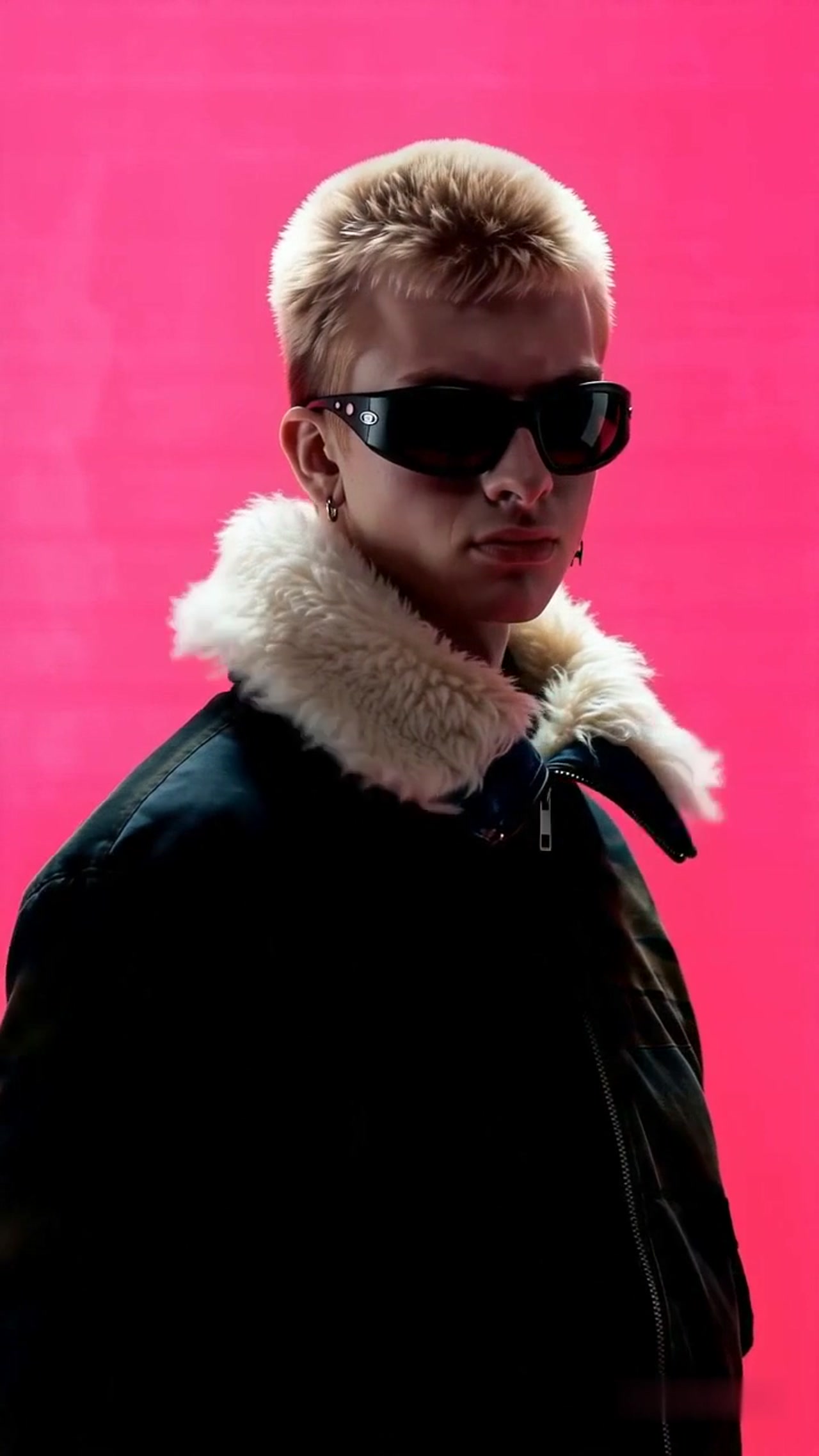 A person in sunglasses and a black jacket against a vivid pink background